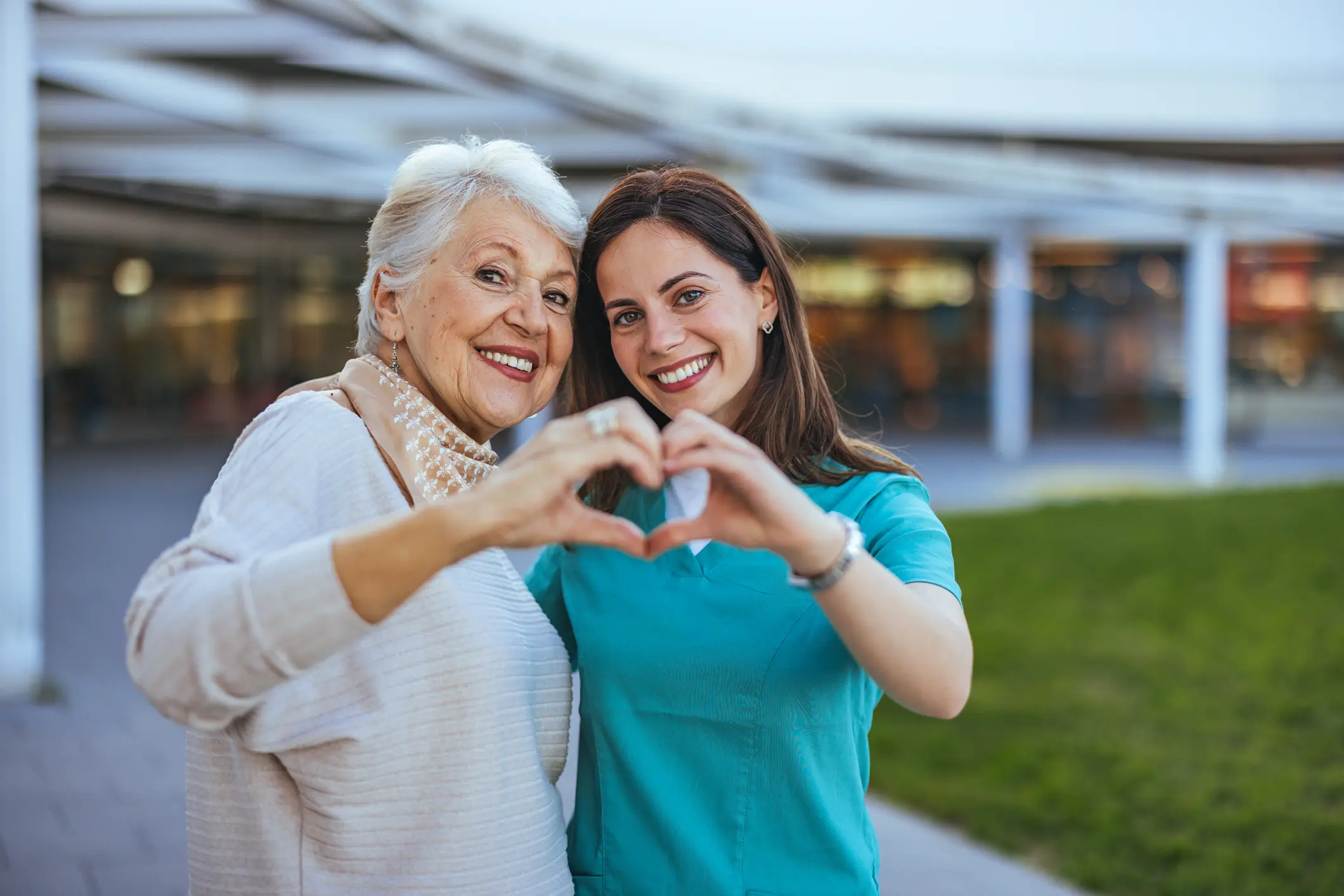 Joyful Senior Woman and Caregiver Making Heart Shape Outdoors Elderly woman and her caregiver happily form a heart shape with their hands, symbolizing care and companionship in an outdoor setting.